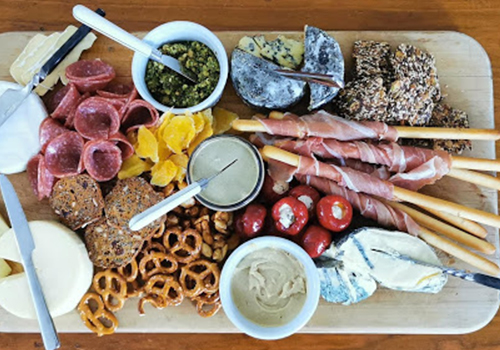 Top-down view of a homely cheese board with blue cheese, havarti and bri, cold meats, pretzels and pesto.