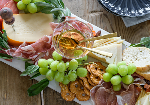Top-down view of a cheese board with green grapes, pretzels, prosciutto, havarti and honey.