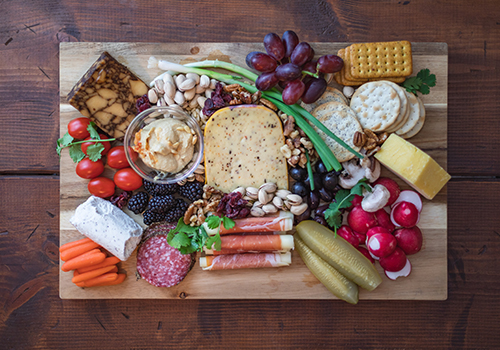 Top-down photo of a cheese board with different types of cheese, pickles, cold meat, fruit, vegetables, nuts and crackers.