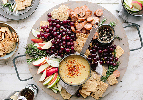 Hot cheese dip on a platter surrounded by grapes, apple slices, crackers and sliced sausage.