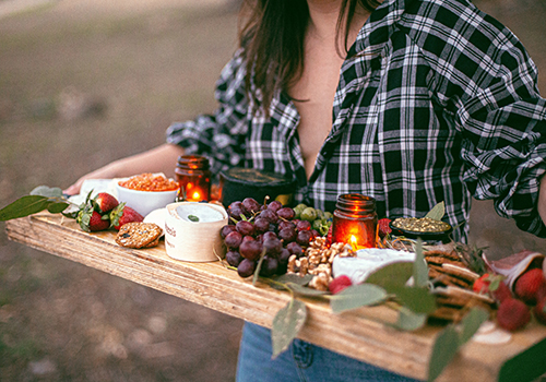 A woman carrying a long board with cheese, tea lights, grapes, strawberries, olives and dips.