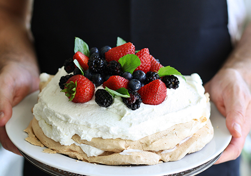 Close up of someone holding a pavlova on a plate with whipped cream, strawberries, blackberries, blueberries and mint.